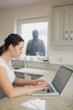 Woman using laptop in kitchen with robber looking at her through windowの写真素材