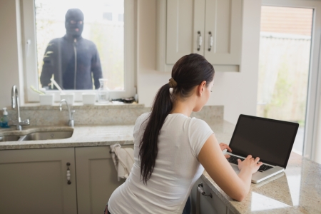 Robber looking at woman in kitchen using laptop through windowの写真素材