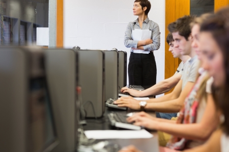 Concentrate students sitting at the computer room while teacher standing at top of class in college looking out windowの写真素材