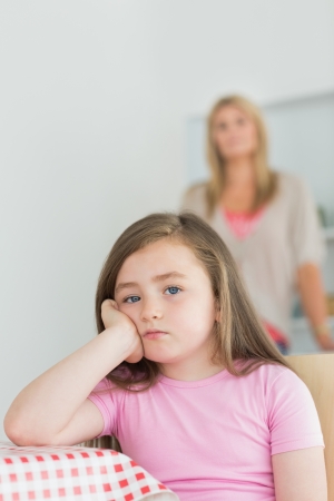 Little girl looking annoyed sitting at kitchen table with mother in backgroundの写真素材