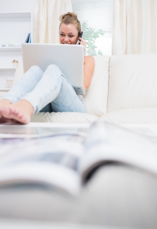 Cheerful woman sitting on the couch while using laptop and calling with mobile phoneの写真素材