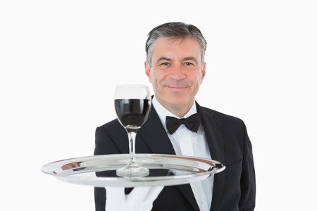 Smiling waiter holding a glass full of wine on a silver tray on white backgroundの写真素材