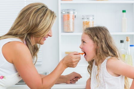 Smiling mother feeding daughter carrot in kitchenの写真素材