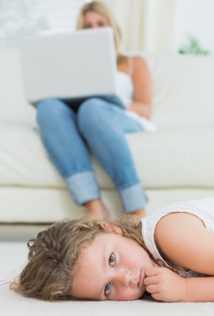 Sad daughter resting on the floor while her mother using notebook on the sofaの写真素材