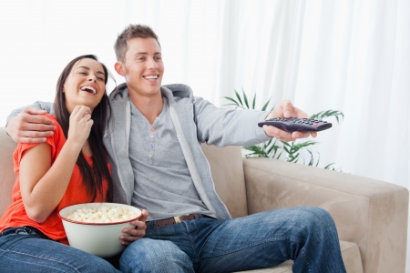 A laughing couple on the couch embracing with popcorn as they watch a tv show の写真素材