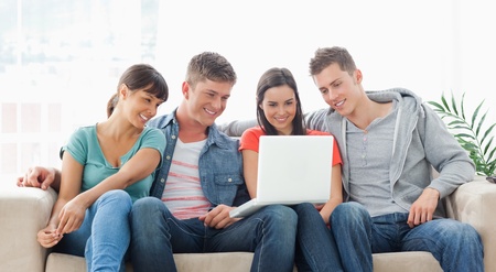 A group of friends sit together on the couch watching the laptop while smiling