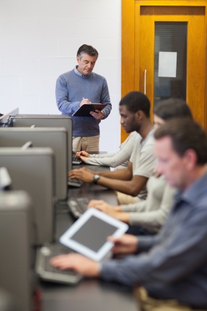 Teacher writing on clipboard during class in computer room in collegeの写真素材