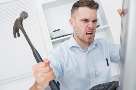 Frustrated young man hitting computer monitor with hammer at officeの写真素材