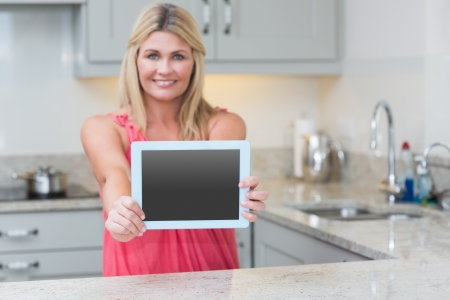 Portrait of casual young woman holding out digital tablet in the kitchen at homeの写真素材