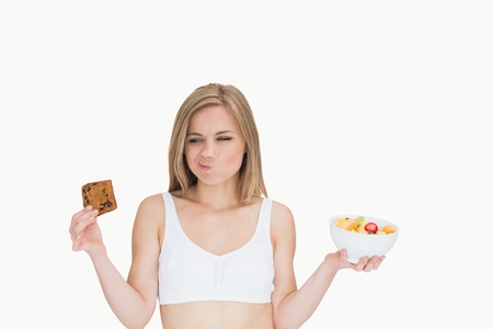 Young woman with fruit bowl making faces as she looks at cookie over white backgroundの写真素材