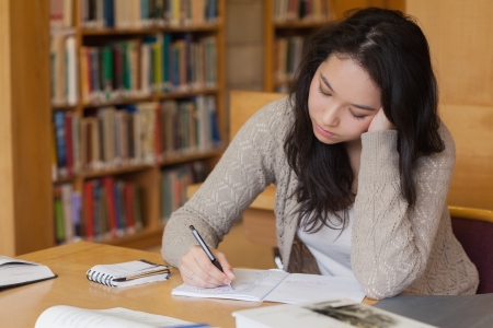Bored student sitting at a desk in a library while taking notes and learning