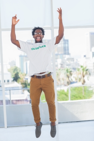 Man jumping with volunteer tshirt then raising his arms in a modern officeの写真素材