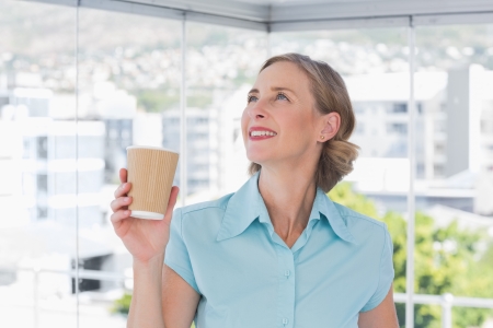 Happy businesswoman holding disposable coffee cup and looking up in bright officeの写真素材