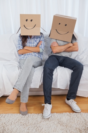 Couple sitting with cardboard boxes with smiley faces on head in living roomの写真素材
