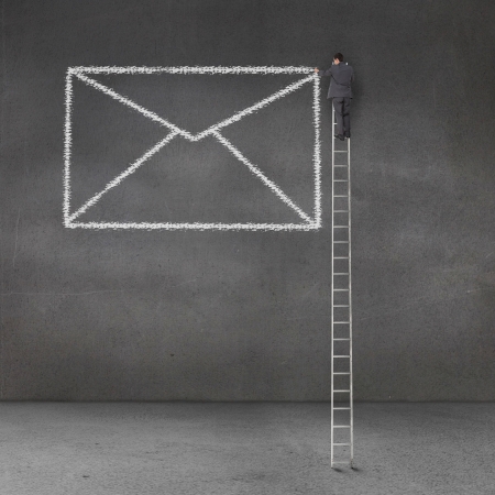 Businessman drawing a giant envelope on a grey wall and standing on a giant ladderの写真素材