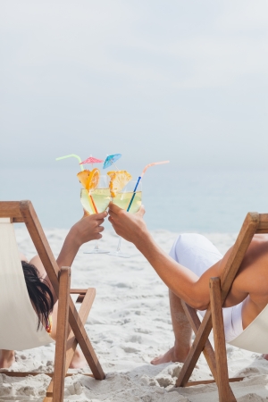 Couple clinking glasses of cocktail on their deck chairs at beachの写真素材
