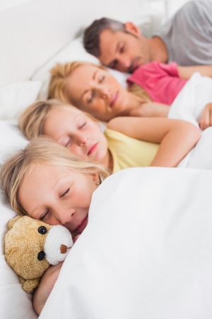 Young girl holding a teddy bear next to her sleeping family in bedの写真素材