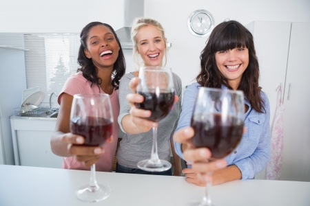 Cheerful friends toasting to the camera with glasses of red wine at home in kitchenの写真素材