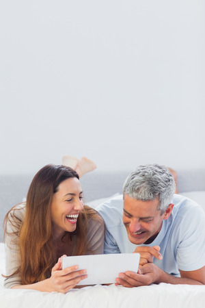 Smiling couple lying on bed using their tablet pc in bedroomの写真素材