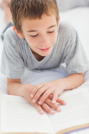 Smiling little boy lying on bed reading book at homeの写真素材