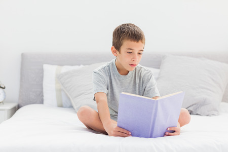 Concentrated little boy sitting on bed reading book at homeの写真素材