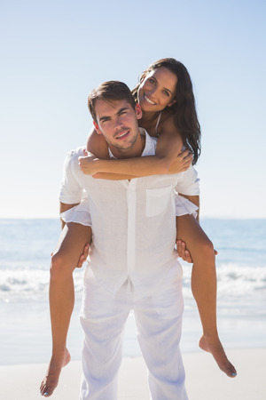 Smiling man giving happy girlfriend a piggy back looking at camera at the beachの写真素材