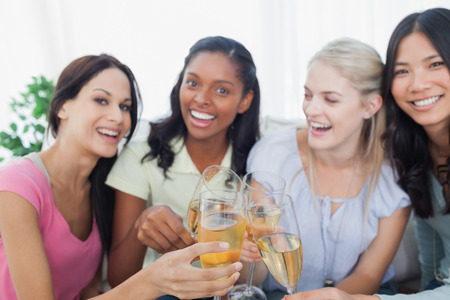 Friends toasting with white wine and smiling at camera at home on couchの写真素材