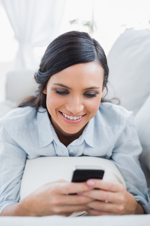 Cheerful pretty woman in her living room lying on the couch sending messagesの写真素材