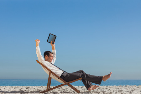 Victorious young businessman on the beach resting on his deck chair holding his tabletの写真素材