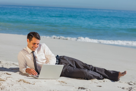 Cheerful young businessman relaxing lying on the sand with his laptopの写真素材