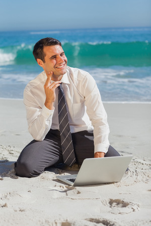 Thoughtful businessman sitting on the sand with his laptop while looking awayの写真素材