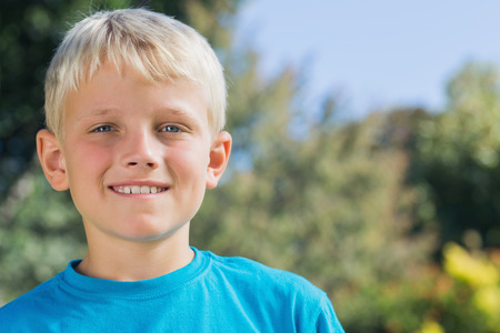 Cute blonde boy smiling at camera in the park with trees behind him