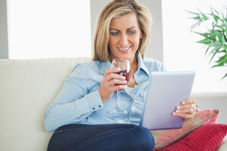 Smiling blonde woman drinking wine and using her tablet pc in a living roomの写真素材