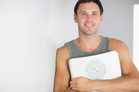Sporty cheerful man leaning against wall holding a scale in bright roomの写真素材