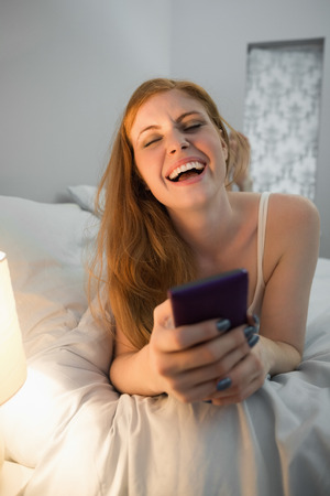Laughing redhead lying on bed sending a text at home in her bedroomの写真素材