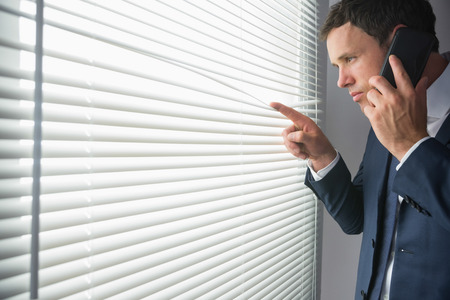 Unsmiling handsome businessman looking through roller blind phoning in dark roomの写真素材