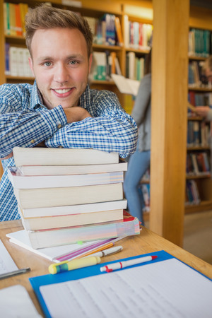Portrait of a smiling male student with stack of books while others in background at the college libraryの写真素材