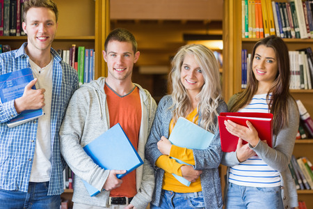 Group portrait of four students with folders standing against bookshelf in the college libraryの写真素材
