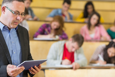 Portrait of an elegant teacher using tablet PC with students sitting at the college lecture hallの写真素材
