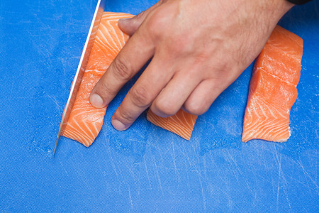 Close up of hand slicing raw salmon with sharp knife on blue cutting board