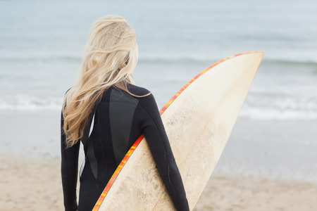 Rear view of a young woman in wet suit holding surfboard at beach