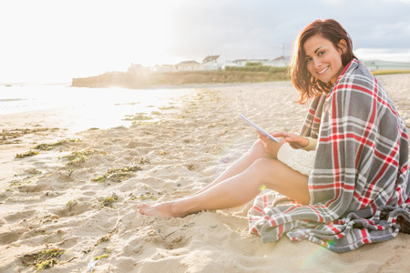 Side view portrait of a young woman covered with blanket using tablet PC at the beachの写真素材