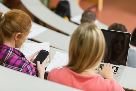 Close-up rear view of two females using laptop and cellphone in the college lecture hallの写真素材