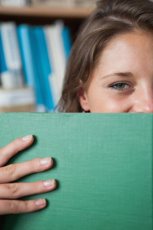 Close-up portrait of a female student holding book in front of her face in the libraryの写真素材