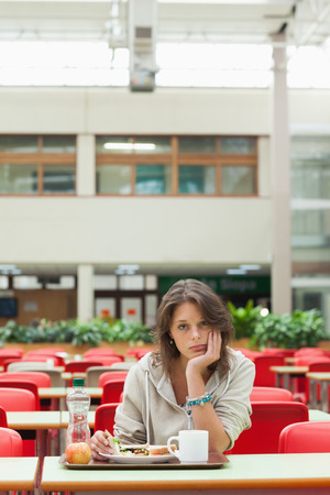 Gloomy female student sitting in the cafeteria with food trayの写真素材