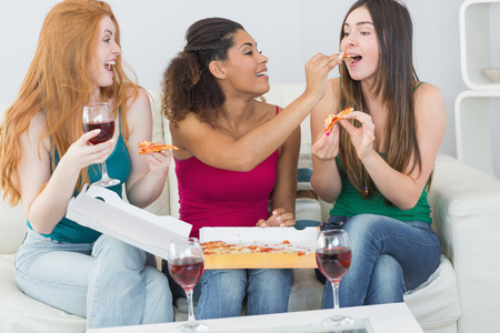 Happy young female friends eating pizza with wine on sofa at homeの写真素材