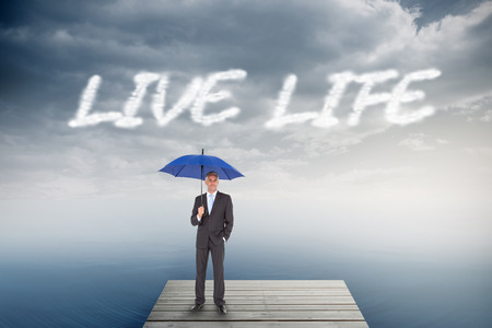 The word live life and businessman smiling at camera and holding blue umbrella against cloudy sky over oceanの写真素材