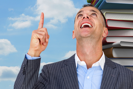 Cheerful businessman pointing upward  against pile of books against skyの写真素材