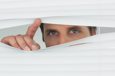 Close-up portrait of a green eyed businessman peeking through blinds in the officeの写真素材