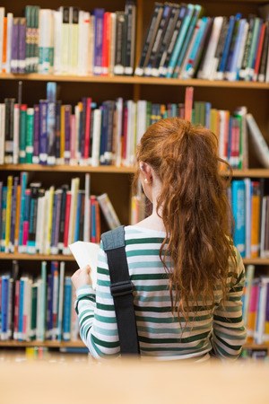 Redhead student reading book from shelf standing in library at the universityの写真素材